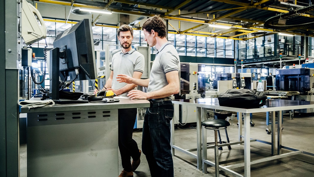 Two male plant managers looking at a desktop monitor in a warehouse workspace.