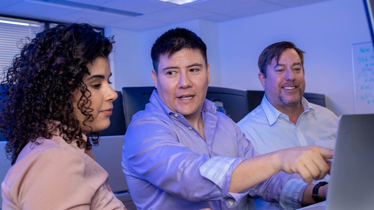 Man sitting in between a woman and another man while pointing at a laptop on a table in front of the group.