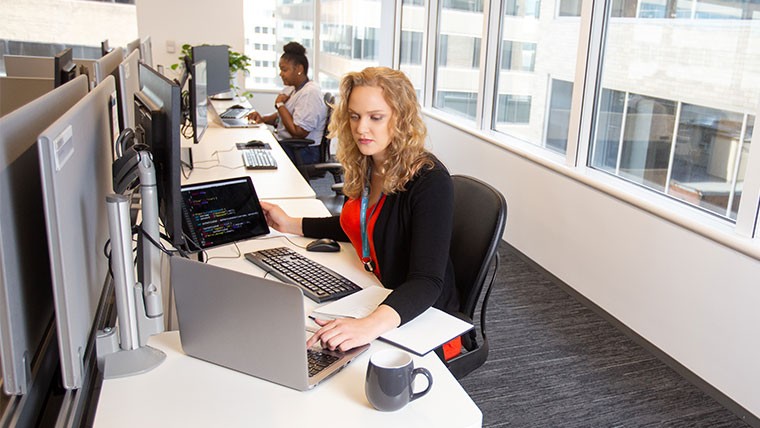 Woman working at a desk on a laptop with a keyboard and monitor in front of her.