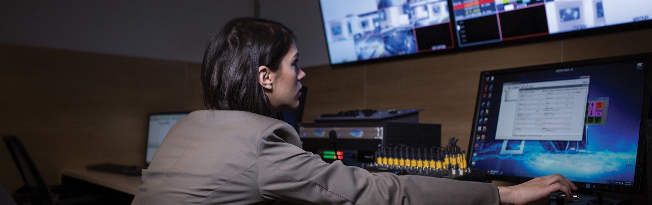 Woman sitting at a desk working on a laptop with several monitors above her on the wall.