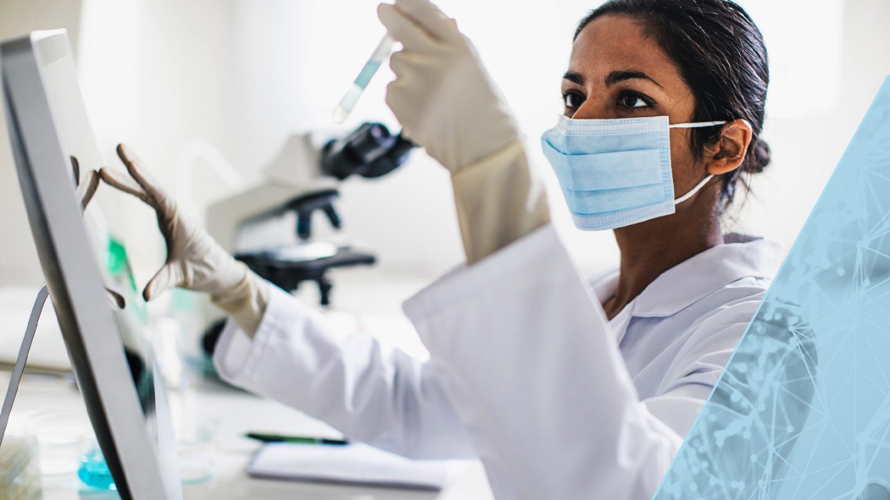 Woman researcher in a mask examining a test tube while also using a touch screen monitor on the desk in front of her.