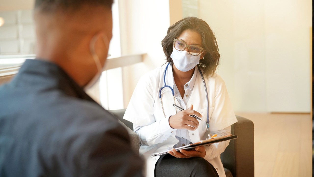 Doctor sitting in a chair holding a chart while talking to a patient.